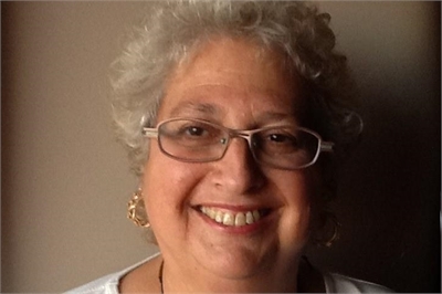 [ai] A smiling woman with curly gray hair wearing glasses and silver earrings, posed against a neutral background. She is dressed in a light top and appears to be in a well-lit setting.